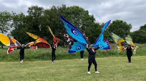 Tom Jackson/BBC Five people wearing black clothes and bright carnivalesque butterfly wings perform a dance on the grass in Nene Park. They wear coloured goggles to look like bug eyes.