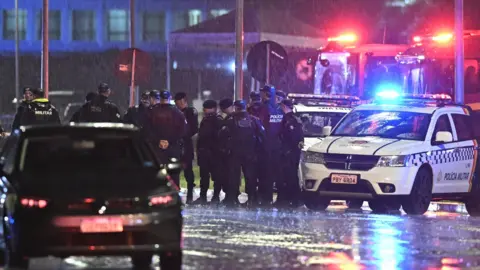 Brazilian police officers stand huddled beside police cars in the capital Brasilia. Heavy rain is falling and puddles can be seen on the ground of the photo. 