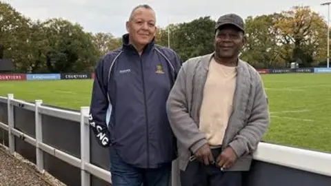 Two men stand side by side smiling into the camera. Both are wearing waterproof style jackets and the man on the right has a dark baseball cap on his head.