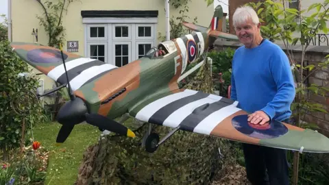 A large model Spitfire plane in a garden. The plane is dark green, white, black and brown with black propellers on its nose. It has an RAF circular logo on the side and camouflage netting underneath it. Standing next to it is Ray, who has white hair and is wearing a blue jumper. His hand is resting on the wing.