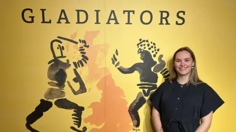 A woman in a black dress stands next to a yellow wall with black outlines of two men fighting with swords and shield with the word "Gladiators" written in black above them.