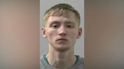 Mugshot of Nathan Kelly. He has short fair hair and a faint blond beard on his chin. He has a number of cuts or spots on his face.