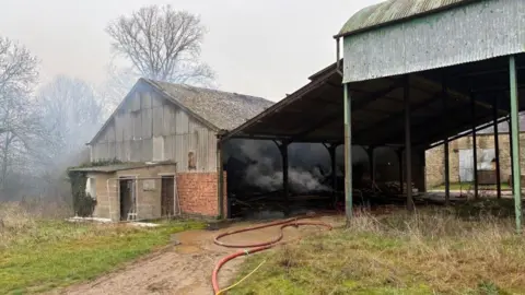 Northamptonshire Fire and Rescue Large red and yellow hoses run across the muddy ground next to a largely metal barn. Grey smoke is billowing out the side and roof of the building. 