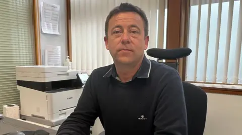 A man sat at a desk with a printer and window behind him.
