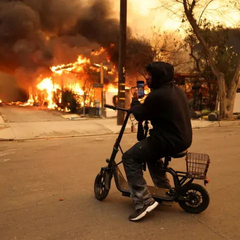 David Swanson/Reuters Seseorang menggunakan ponselnya untuk mengambil gambar gedung yang terbakar sambil duduk di atas skuter yang tidak bergerak dengan tudung terpasang di Eaton Fire di Altadena pada hari Rabu.