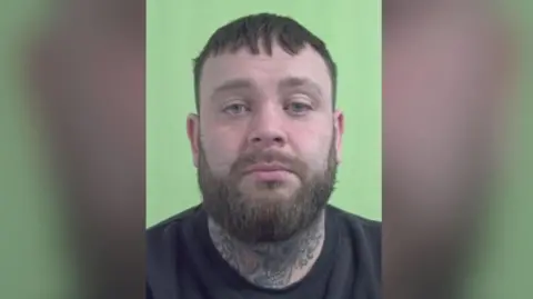 IOM Police A mugshot of John Laird, a man with brown hair and a beard. He is wearing a black top and his neck is covered in tattoos.
