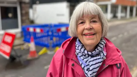 George Carden / BBC Hilary Fritche standing in front of the sinkhole wearing a bright pink jacket and a blue and white striped scarf. She has bright pink lipstick on and bobbed white hair. She is smiling