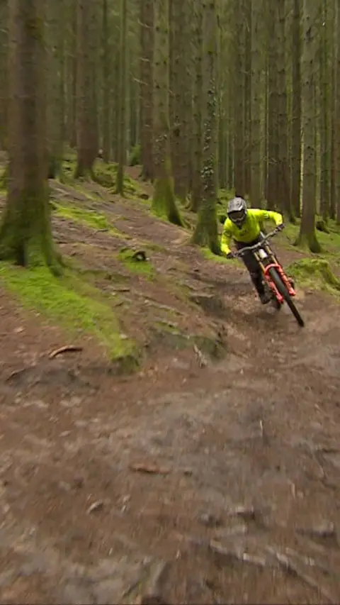 Teenager cycling on a dirt track with trees around him and a bright hi-vis jacket on.