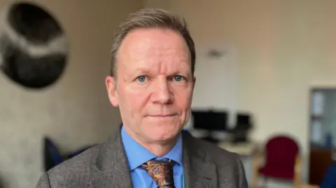 Joe Teape is wearing a blue shirt, a brown patterned tie and a brown suit jacket and is in an office with the background blurred