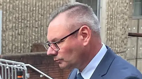 Richard Crisp leaves court. He has grey hair, combed over to one side and wears glasses. He does not look at the camera and wears a blue suit and tie.