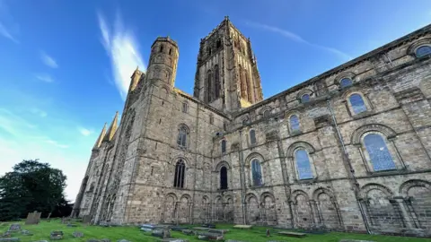 View looking up at the side of Durham Cathedral. The stone building, in Romanesque architectural style, has a high rectangular tower and smaller round ones in front. The windows are high and arched. Gravestone and tombs can be seen at ground level underneath a sunny sky.