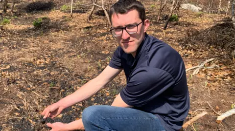 A man is kneeling on burnt ground and holding some blackened soil in his hands. He has brown hair and wears glasses with black frames. He is wearing a navy coloured t-shirt and blue jeans.