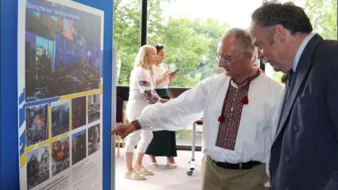 North Yorkshire Council Two men standing in front of a board surrounded by a blue and yellow border covered in photographs of Ukraine and Ukrainian refugees