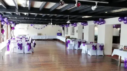 Perry St Social Club A large wooden-floored hall set up for a functon with tables and chairs set out with white table cloths and chair covers and purple ribbons. There are bunches of purple balloons  around the room and black wooden beams across the ceiling