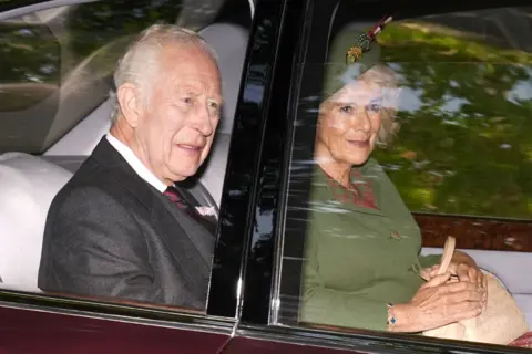 PA Media Charles and Camilla, who is wearing a green outfit and hat, in the back seat of a car looking out the window