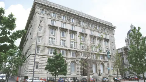 Image shows Liverpool City Council's Cunard Building headquarters. It is a neo-classical five-storey building surrounded by young trees. 