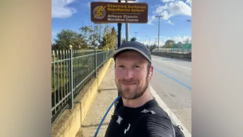 Mike Jones Mike Jones at the side of the road talking a selfie in front of the sign for the Athens Classic Marathon Course in Greece in December 2024.