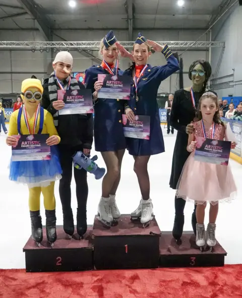 Kali Communications Six ice skaters stand on a podium. On the left, in the second place position is a child dressed in yellow as a Minion and an older child to her right wears a Gru costume, from the film Despicable Me. In the middle, in the first place position, are two women in air hostess uniforms. On the right, in the third place position, are a woman and child dressed as Elphaba and Glinda from Wicked.