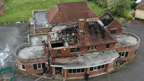 Ian Bateman An empty red brick pub, that has been damaged by fire. The roof is severely damaged. One part of the building is round, with different sections to the building. There is a grass area behind the pub and another building to the right. 