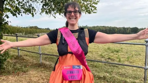 Supplied Jennifer Gane is standing in a field and is wearing an orange ballgown and a pink sash. She also has a Great North Run competitor number pinned to her front which reads: "Jennifer 54351". She is smiling broadly and has her arms outstretched.
