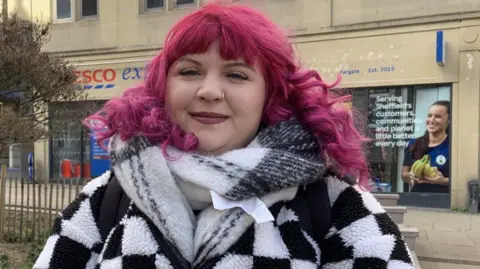 Lucy Ashton/BBC A woman with curly pink hair stands in Sheffield city centre. She is wearing a black and white checked coat