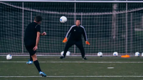 A man in a black kit shoots a football towards the goal with a man in a black kit in goal attempting to save the shot. There are a number of other white footballs in the back of the net. 