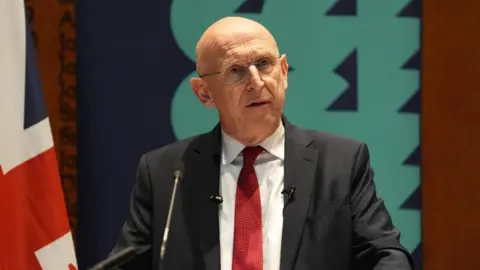 John Healey wearing a dark suit and red tie standing before a microphone