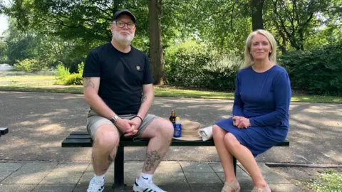 A man and a woman sat on a bench looking at the camera. He s wearing light shorts and a black t-shirt and a cap and has glasses. She is wearing a blue dress and has long blonde hair. In between them is a hat and two sunscreen bottles. They are in a park. 