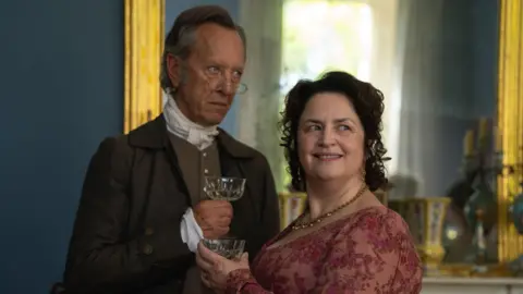 BBC/Bad Wolf/BritBox A man and woman in Regency period costume stand together in a still from The Other Bennet Sister. Richard E Grant is peering over his glasses while standing next to Ruth Jones. Both are holding a glass of what appears to be wine. A gold gilted mirror and fireplace can be seen behind them.