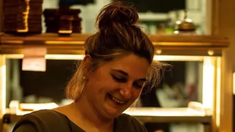 Domaine 16 Stephanie Ronssin smiling. She is standing in a restaurant with her hair in a bun.