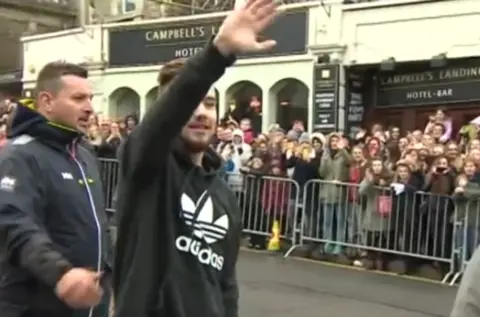 One Direction member Liam Payne waves to fans in Clevedon as the band visited to film a music video in 2014. Rows of fans can be seen behind crash barriers. Payne is wearing a black hoodie with a white Adidas logo on it