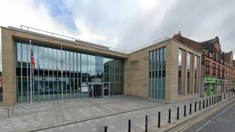 Google Cumberland Council's headquarters in Carlisle: a two-storey building with high windows. There is a small square and two flags at the front, including the Union Jack. 