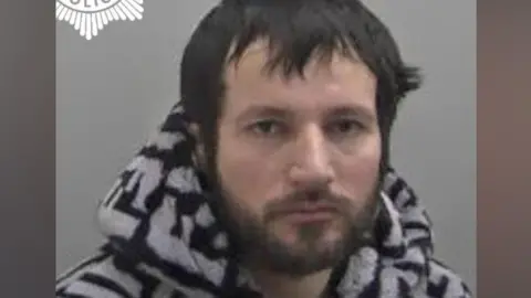 A police mugshot of a man wearing a black and white patterned jumper. He has black hair and a black beard. He is positioned against a light-coloured backdrop.