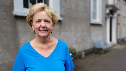 Janet is wearing a blue top and standing in front of the church