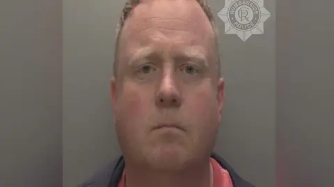 Humberside Police Robert Bush with short, light brown hair is shown in a straight‑on mugshot photograph. He has a neutral expression and is wearing a dark jacket over a red shirt. The background is plain and grey, and a Humberside Police badge watermark appears in the upper right corner of the image.
