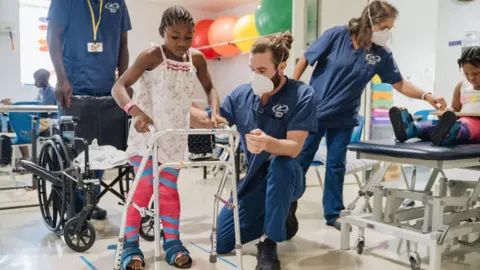 Mercy Ships Fudia is in a physical therapy room after her surgery. There is a physiotherapist standing behind her holding a wheelchair, she is standing upright with another physiotherapist kneeling by her side. Fudia wears her hair in braids, a thin strapped white nightie with red and pink floral print. She has neon pink casts on both legs, with blue taping and velcro strapped boots on her feet. Fudia is holding onto a walking frame. The male physical therapist next to her is supporting Fudia with his hand at her back, he has long brown hair tied into a bun and wears a medical face mask. There are other children in the background receiving support from therapists too.