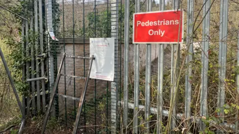 Carmelo Garcia A close off bridge with a red 'pedestrians only' sign on metal fencing. There is a small ladder leaning up against a metal railing and lots of overgrown plants round it. 