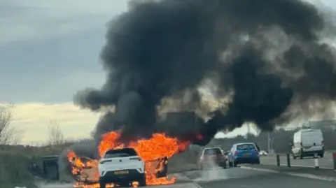 A white car engulfed in orange flames with a large black cloud of smoke rising in the air. The car is parked in a lay-by while other cars are still driving on the road.