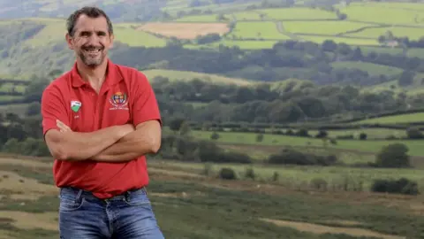 NFU Cymru Garry wearing a red polo shirt and blue jeans with short dark hair and a beard stands smiling with his arms crossed. Behind him is a wide landscape of a farm