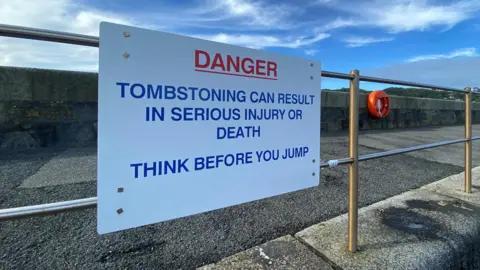 Guernsey Coastguard A white sign on a railing which says "Danger, tombstoning can result in serious injury or death, think before you jump".
