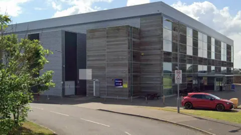 Google A sleek, contemporary building with wide glass panels and metal cladding sits beside a quiet road. A red car is parked out front, framed by green trees and a bright, partly cloudy sky.