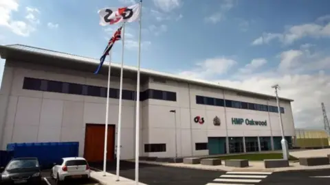 A two storey white building with the word HMP Oakwood in blue signage. There are three flag poles with a G4S flag, a Union Jack flag and blue flag while two cars, one black and another white, are parked outside the prison. 