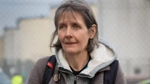 Dr Sarah Benn at a protest. She has bobbed grey hair and is wearing a hoodie with a sheepskin-stlye hood and a backpack. The background is blurred. 