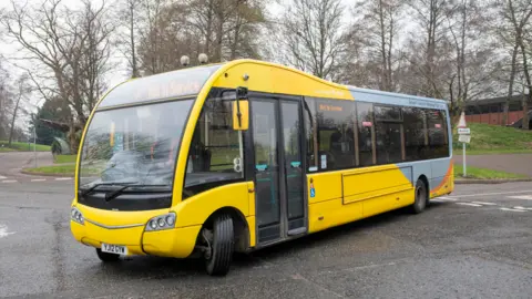 A modern, yellow, medium sized bus shown driving around a corner on quiet roads