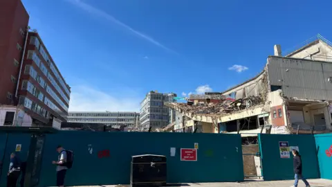 Owen Sennitt/BBC A man walks along a street looking towards the Anglia Square demolition site, with a partially demolished former cinema building in the background