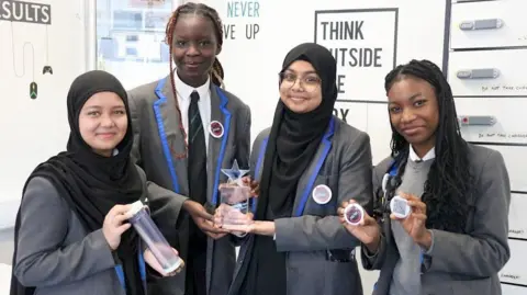 [L-R - Tajkia (14), Neveah (15), Sabriha (15), Daniela (15) ] Pupils pictured in grey school uniform hold a trophy and badges