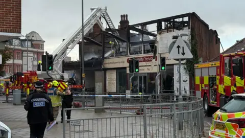 Elliot Deady/BBC Fire crews and police on the scene this morning. Hoses are being used to spray down a brick building with missing windows, a missing roof and charred walls. The sign on the front of the building is pale yellow and reads 'easy mobility services'.