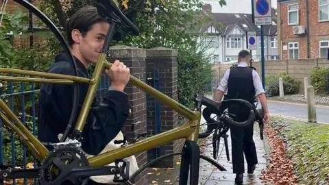 Alasdair Bayman Alasdair is holding his bike outside, it is mustard yellow in colour and does not have any wheels. There is a policeman holding another bike ahead.