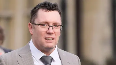 Andrew Matthews/PA Wire A man in a white shirt and tie, wearing a light grey suit and glasses. He has short brown hair and grey stubble.