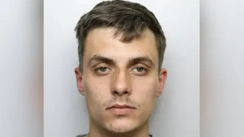 Custody shot of Regan Kemp. He has short brown hair, has a serious expression and is stood in front of a grey background. 